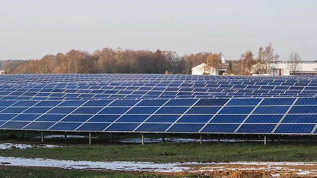 Solpaneler i långa rader på öppen mark, i bakgrunden industribyggnader.