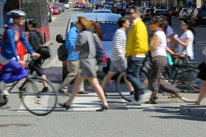 Fotografi på ett övergångsställe. Många människor och cyklister passerar framför biltrafiken.