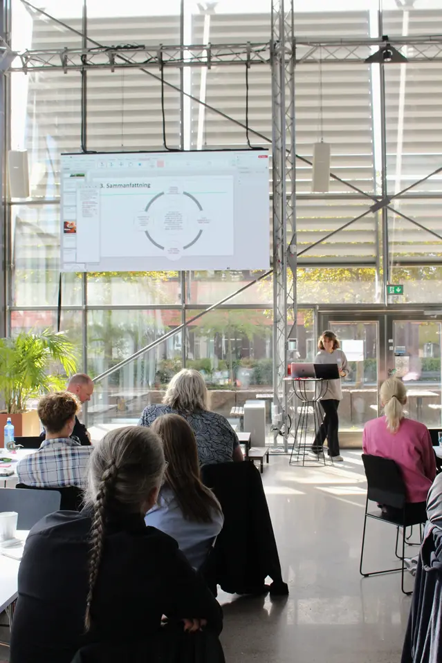 Ryggarna på sittande projektdeltagare syns framför en tjej håller presentation längst fram i ett rum.