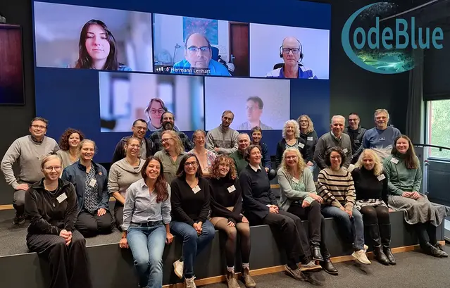 A group of people on a stage and some people are seen on the screen behind them, particiating online.