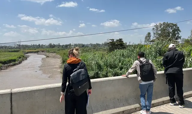 En brunfärgad flod rinner sakta förbi där tre personer står på bron och tittar dedåt.
